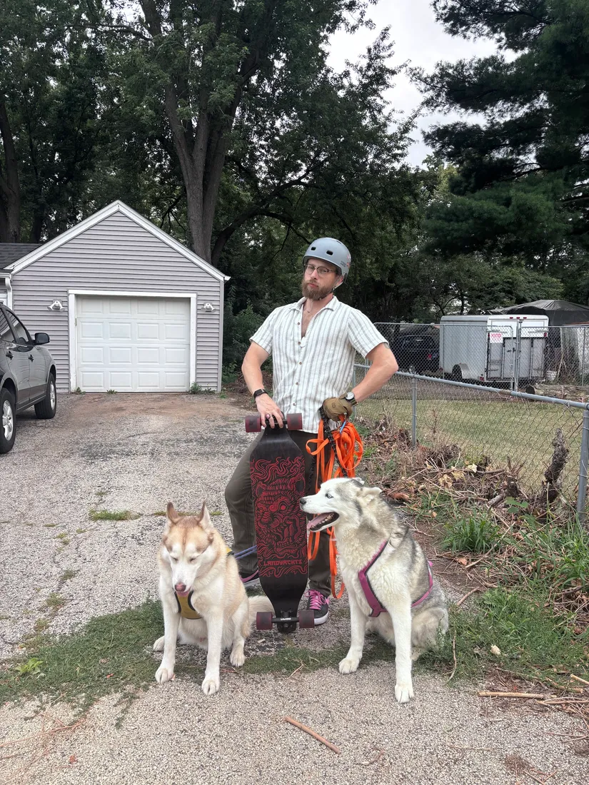 Joshua Herrington in a helmet with a longboard, flanked by two Siberian Huskies