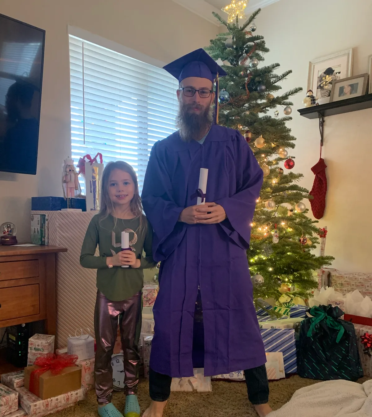 Joshua Herrington in cap and gown at home, beside his daughter holding her own rolled diploma